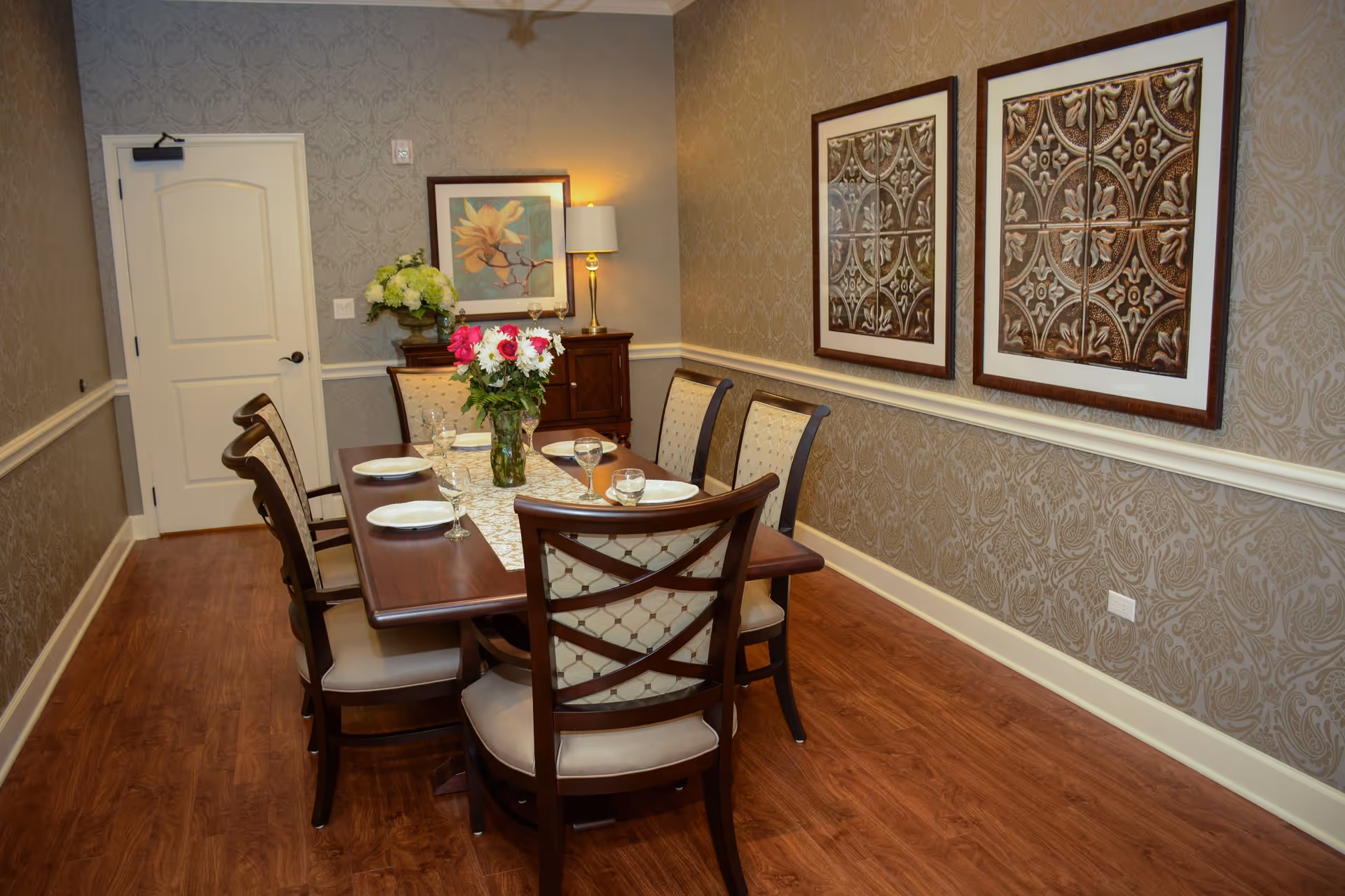 A formal dining room with a wooden dining table set for six people. The table has a floral centerpiece and is surrounded by six upholstered chairs with wooden frames. The room features patterned wallpaper, two framed decorative artworks on the wall, a sideboard with a lamp and flower arrangement, and a closed white door. The floor is wooden.