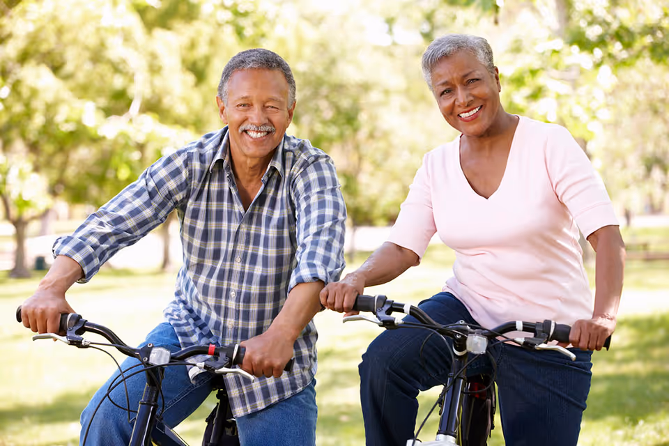 An elderly man and woman smiling and riding bicycles outdoors in a park with green trees in the background.