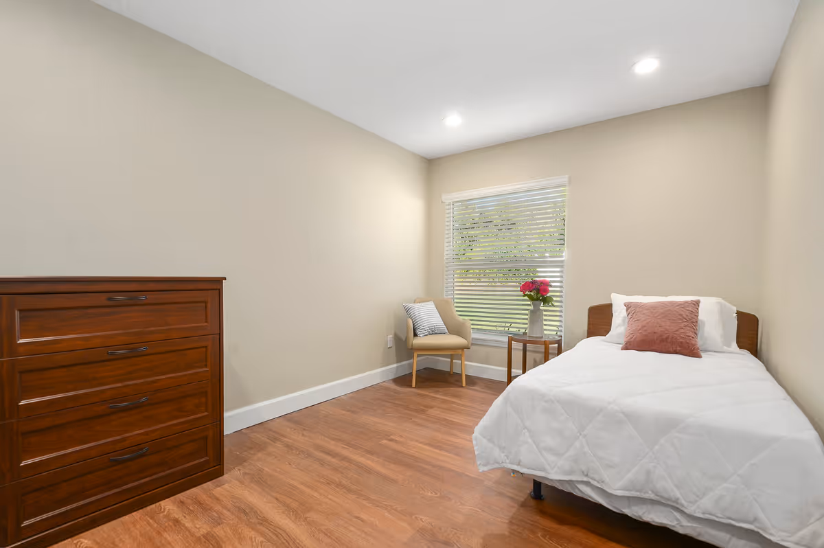 A simple, clean bedroom with a single bed covered in a white quilt and a pink pillow. Next to the bed is a small round wooden table with a vase of pink flowers. A beige armchair with a striped pillow is placed near a window with white blinds, and a wooden dresser with four drawers is against the opposite wall. The room has wooden flooring and beige walls with recessed ceiling lights.