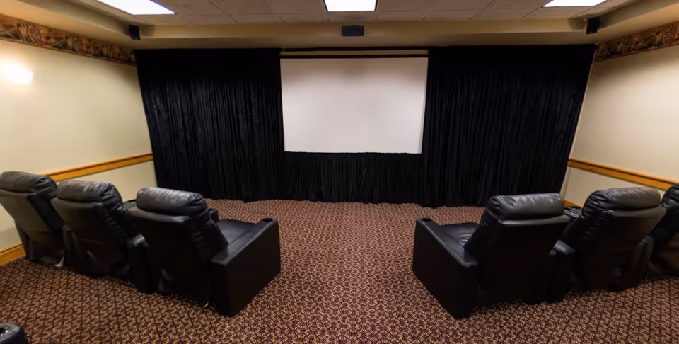 Small screening room with a projection screen and black curtains facing two rows of black leather recliners.