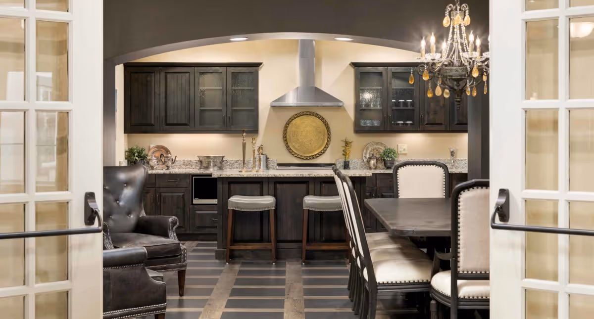 View through glass-paneled double doors into a kitchen and dining area featuring dark wood cabinets, granite countertops, a stainless steel range hood, a kitchen island with two stools, a dining table with upholstered chairs, a leather armchair, and a chandelier overhead.