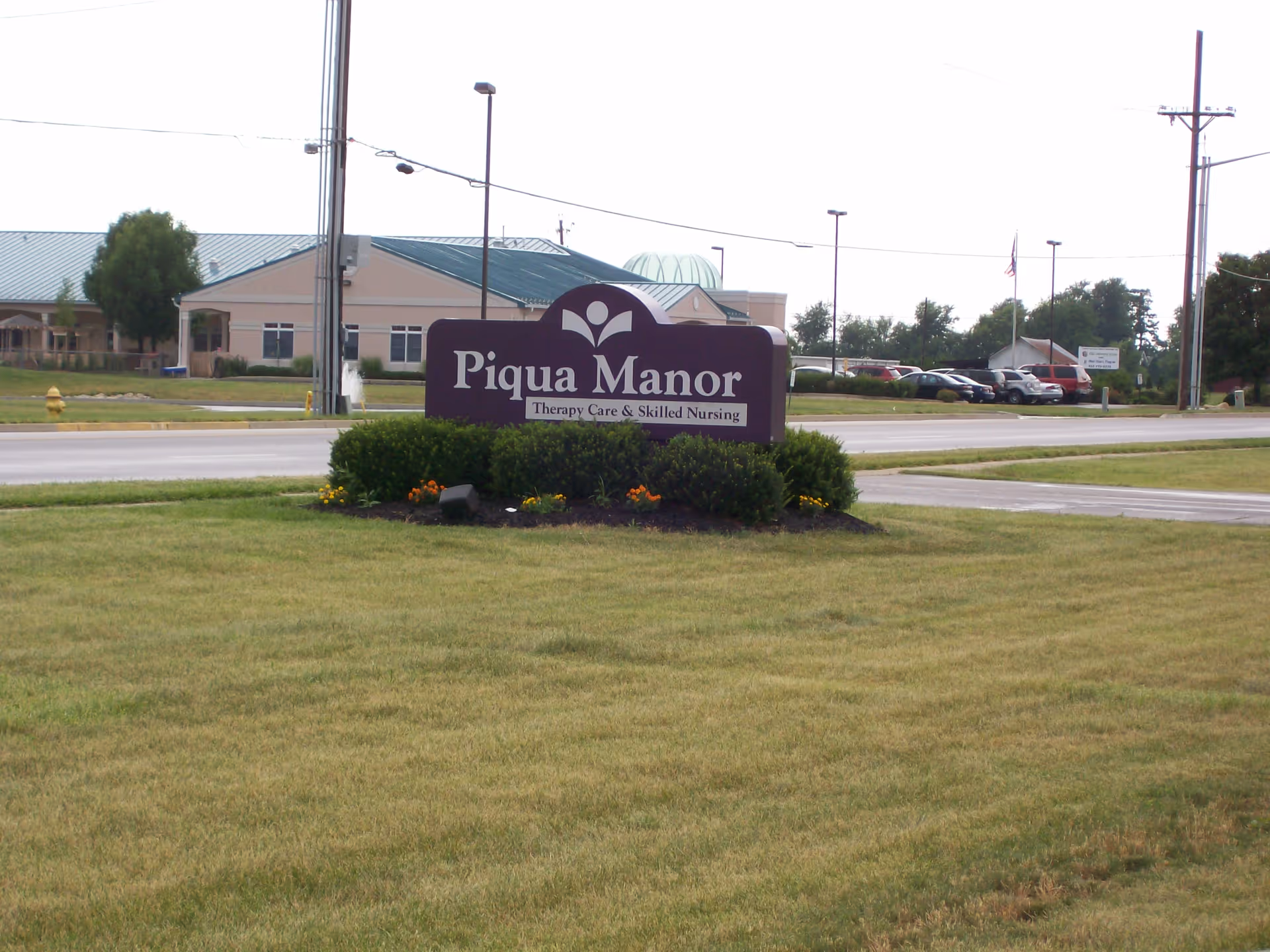 Outdoor view of the Piqua Manor sign on a grassy area with bushes and flowers, with the facility building and parking lot visible in the background across the street.