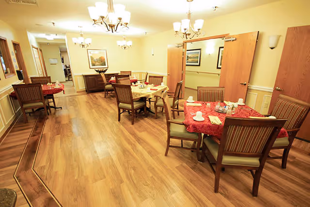Interior view of a dining room in a senior living facility with wooden floors, several tables covered with red tablecloths, chairs arranged around the tables, and chandeliers hanging from the ceiling. The walls are light-colored with framed artwork and wooden doors.