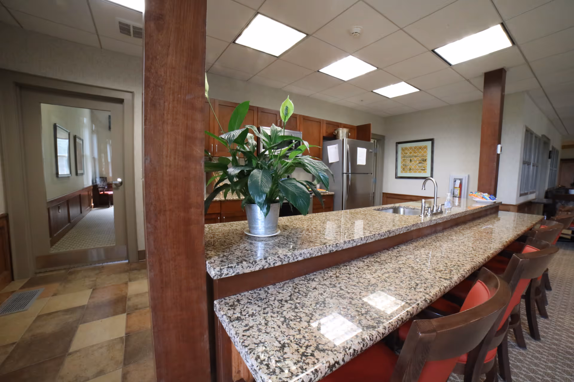 Granite-topped communal kitchen/bar area with a potted plant, sink, refrigerator and several bar stools.