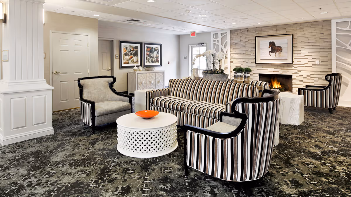 A cozy living room area in a senior living facility featuring a striped sofa and matching armchairs arranged around a white round coffee table with an orange bowl on top. The room has a patterned carpet, a fireplace with a horse painting above it, decorative plants, and framed artwork on the walls. The space is well-lit with ceiling lights and has a neutral color palette with white and beige tones.