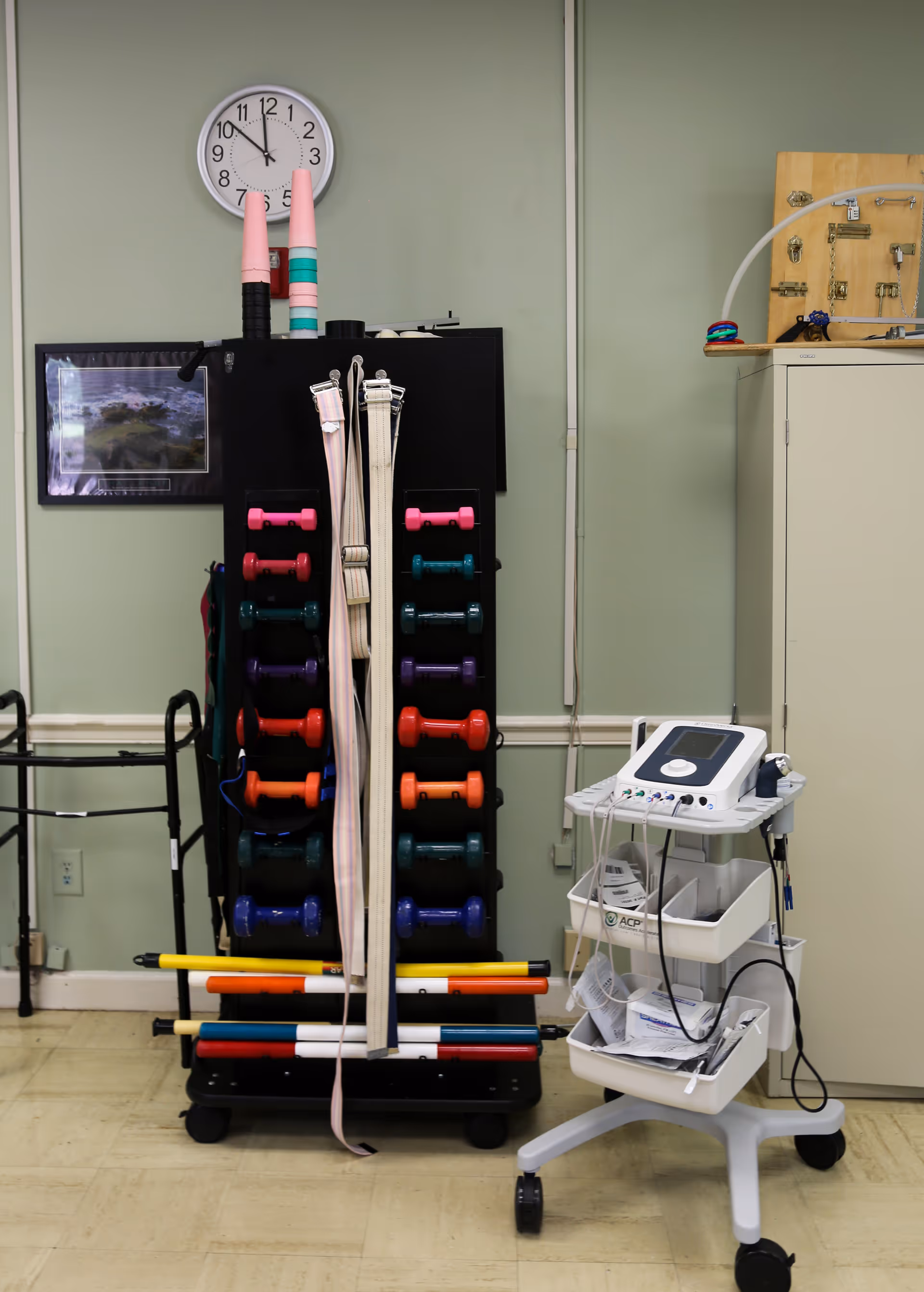 A therapy or rehabilitation room with a rack holding various colorful dumbbells, exercise belts, and rods. To the right, there is a medical device on a wheeled cart with cables and papers. A clock on the wall shows the time as 11:55. The walls are painted light green, and there is a framed picture and a cabinet in the background.