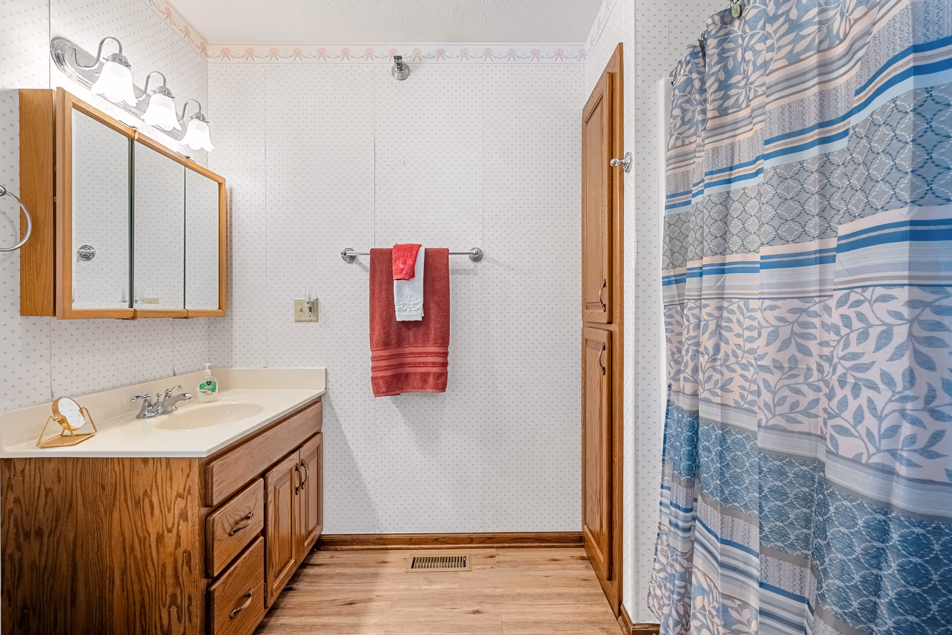Bathroom with a wood vanity and mirrored cabinet, a red towel on a wall rack, and a blue patterned shower curtain.
