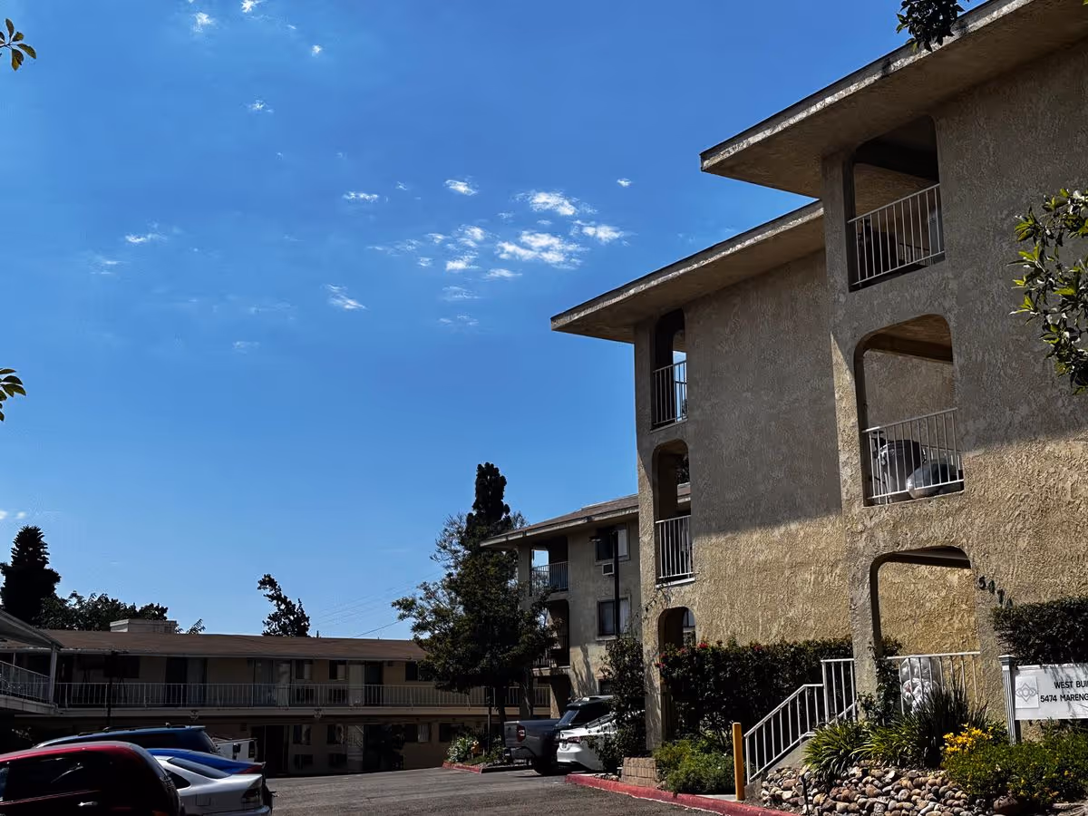 Exterior of a multi-story senior living apartment building with balconies, landscaping and parked cars under a blue sky.