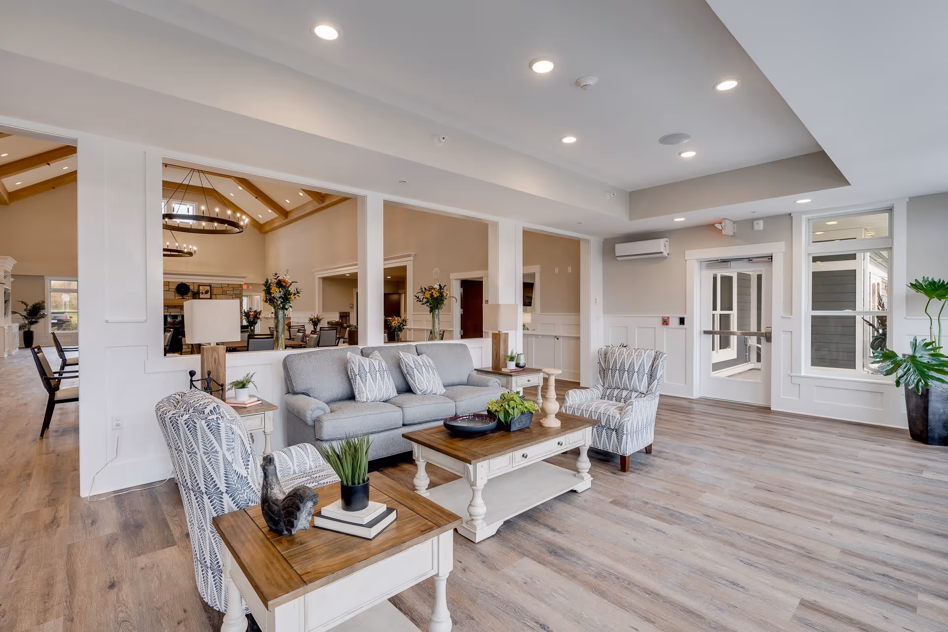 A bright and spacious living room area in a senior living facility named Rivers of Life. The room features a gray sofa with patterned pillows, two patterned armchairs, a wooden coffee table with decorative items, and side tables with lamps and plants. The floor is light wood, and large windows and glass doors allow natural light to enter. In the background, a dining area with tables, chairs, and floral centerpieces is visible through large interior windows. The ceiling has recessed lighting and a modern design.