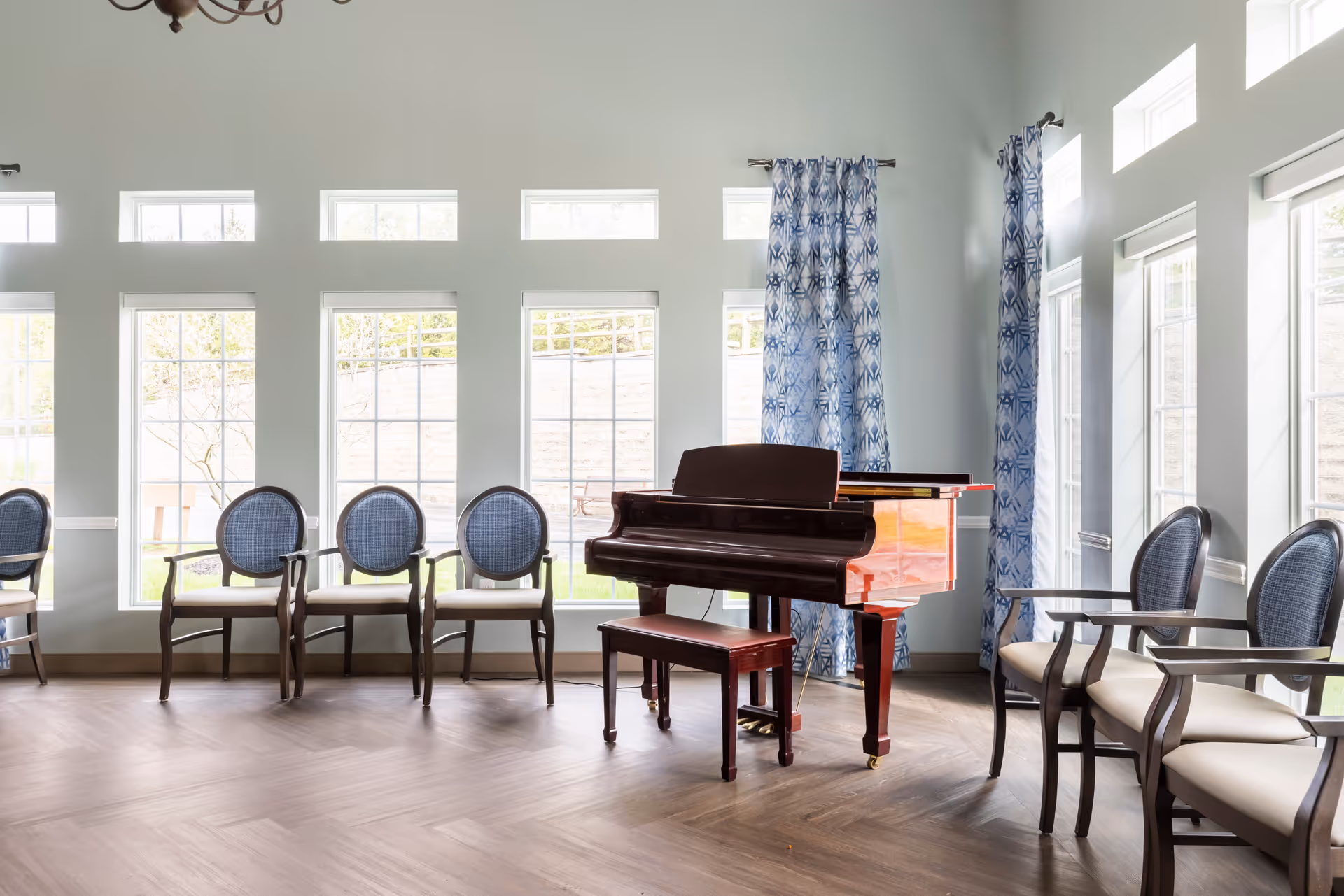 A bright room with large windows and blue patterned curtains, featuring a dark wooden grand piano with a matching bench in the center. Surrounding the piano are several wooden chairs with blue cushioned backs and light-colored seats arranged along the walls.