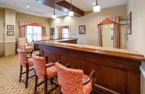 Interior view of a common area in an assisted living facility featuring a wooden bar counter with three upholstered bar stools in red patterned fabric. Behind the bar are two windows with red valances and beige walls adorned with framed pictures. The ceiling has wooden beams and hanging pendant lights, and there is a patterned carpet on the floor.