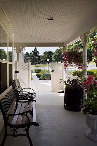 Covered porch area with a wooden bench and a cushioned chair on the left side, hanging flower baskets and potted plants on the right, white decorative columns, and a view of a garden and pathway leading outside through a white gate.