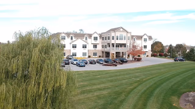 A large three-story senior living facility building with a beige and stone exterior, surrounded by a well-maintained lawn and trees. Several cars are parked in front of the building under a partly cloudy sky.