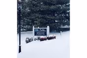 Snow-covered ground and bushes with a sign for Our Lady of the Valley Nursing Center and Rehabilitation Center surrounded by tall evergreen trees in the background.
