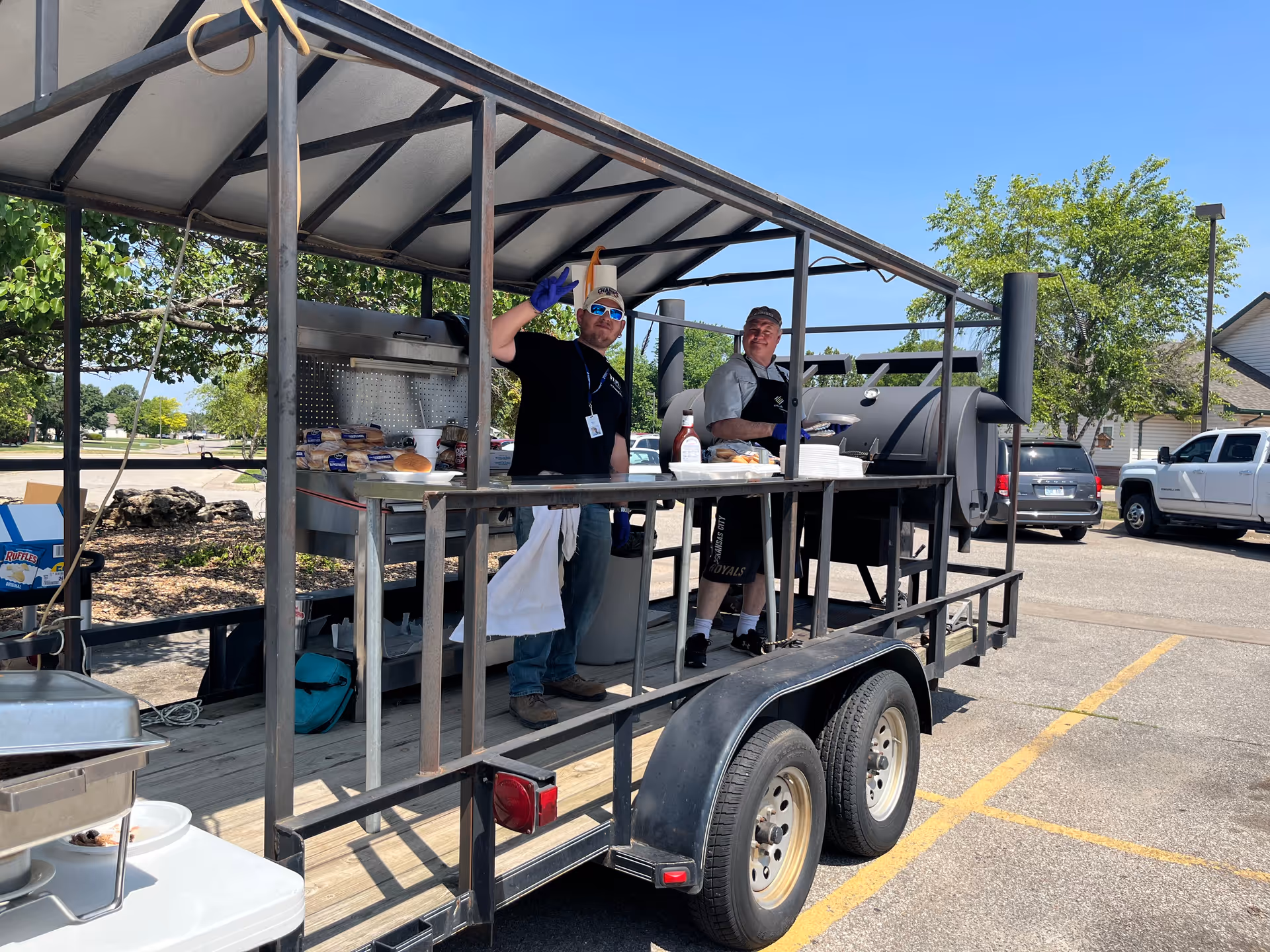 Two men cooking at a large barbecue smoker mounted on a trailer under a canopy in a parking lot.