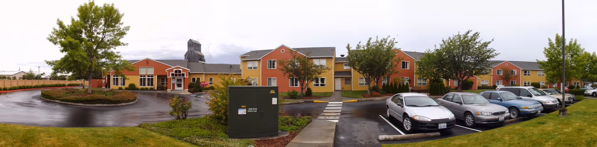 Front exterior of Suncrest Village Retirement Community showing a two-story yellow-and-red building with an entrance drive, lawn, trees, and parked cars.