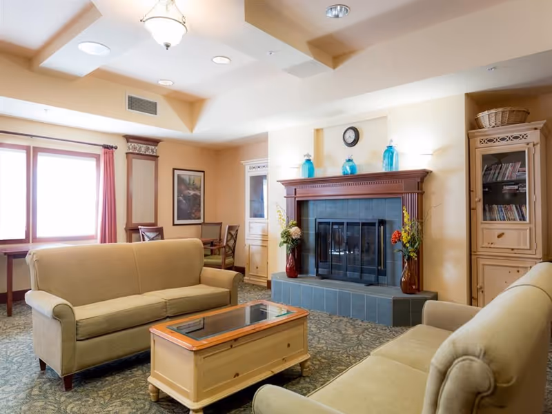 A cozy living room with two beige sofas facing each other, a wooden coffee table with a glass top in the center, and a fireplace with a wooden mantle decorated with blue glass bottles and flower vases on either side. There is a cabinet with shelves filled with books and DVDs on the right, and a window with red curtains on the left letting in natural light. The room has a patterned carpet and soft yellow walls.