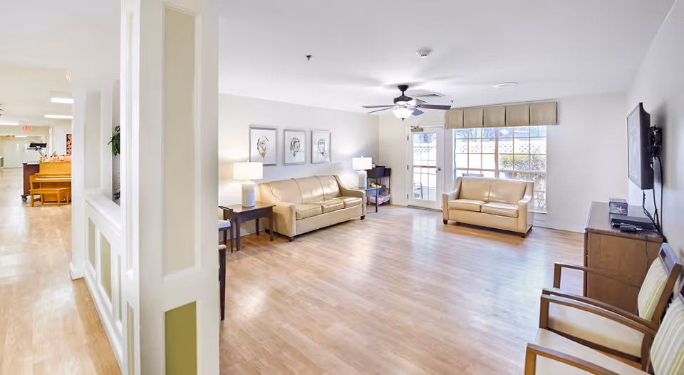 A bright and spacious living room area in a senior living facility with light wood flooring, beige leather sofas, two side tables with lamps, a ceiling fan with light, a wall-mounted TV, and large windows with a door leading outside. The room is decorated with three framed artworks on the wall and has additional seating with two chairs near the TV.