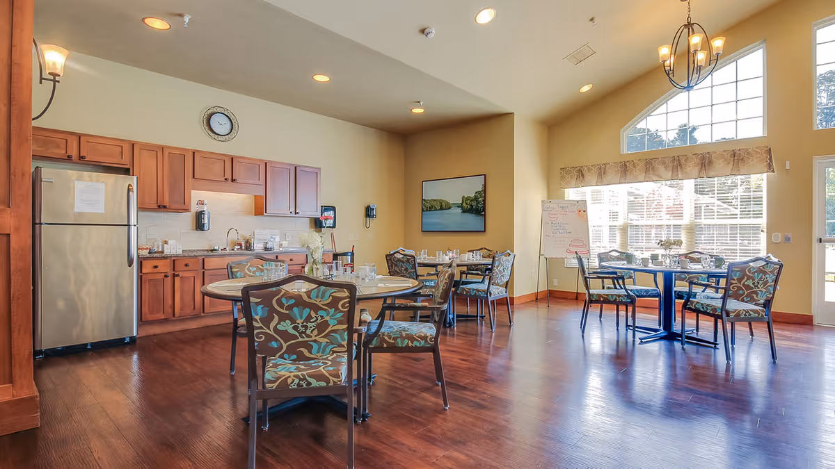 A bright dining area in an assisted living facility featuring wooden floors, round tables with floral upholstered chairs, a kitchen area with wooden cabinets and a stainless steel refrigerator, large windows letting in natural light, and a wall clock above the kitchen cabinets.