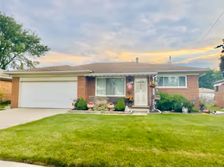 Single-story brick house with a two-car garage, a well-maintained green lawn, and a partly cloudy sky at sunset.