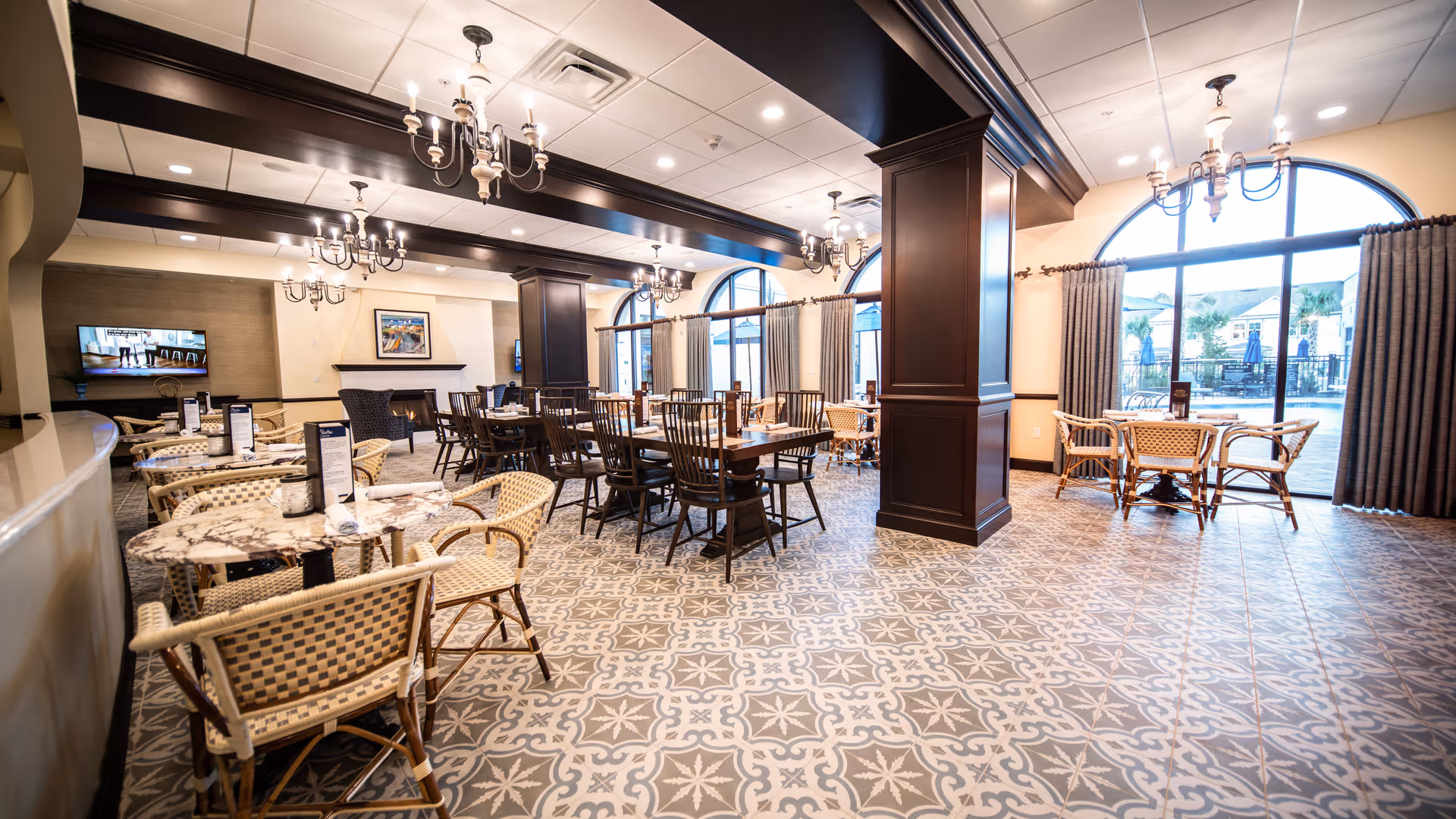 Spacious dining room with multiple tables and chairs arranged neatly. The room features patterned tile flooring, large arched windows with curtains allowing natural light, dark wooden beams and columns, chandeliers hanging from the ceiling, and a fireplace with a mounted TV above it.