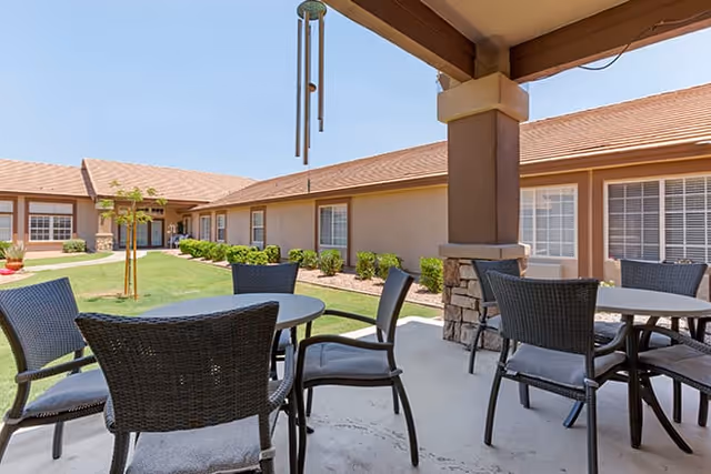Covered patio with round tables and wicker chairs overlooking a grassy courtyard and a single-story senior living building.