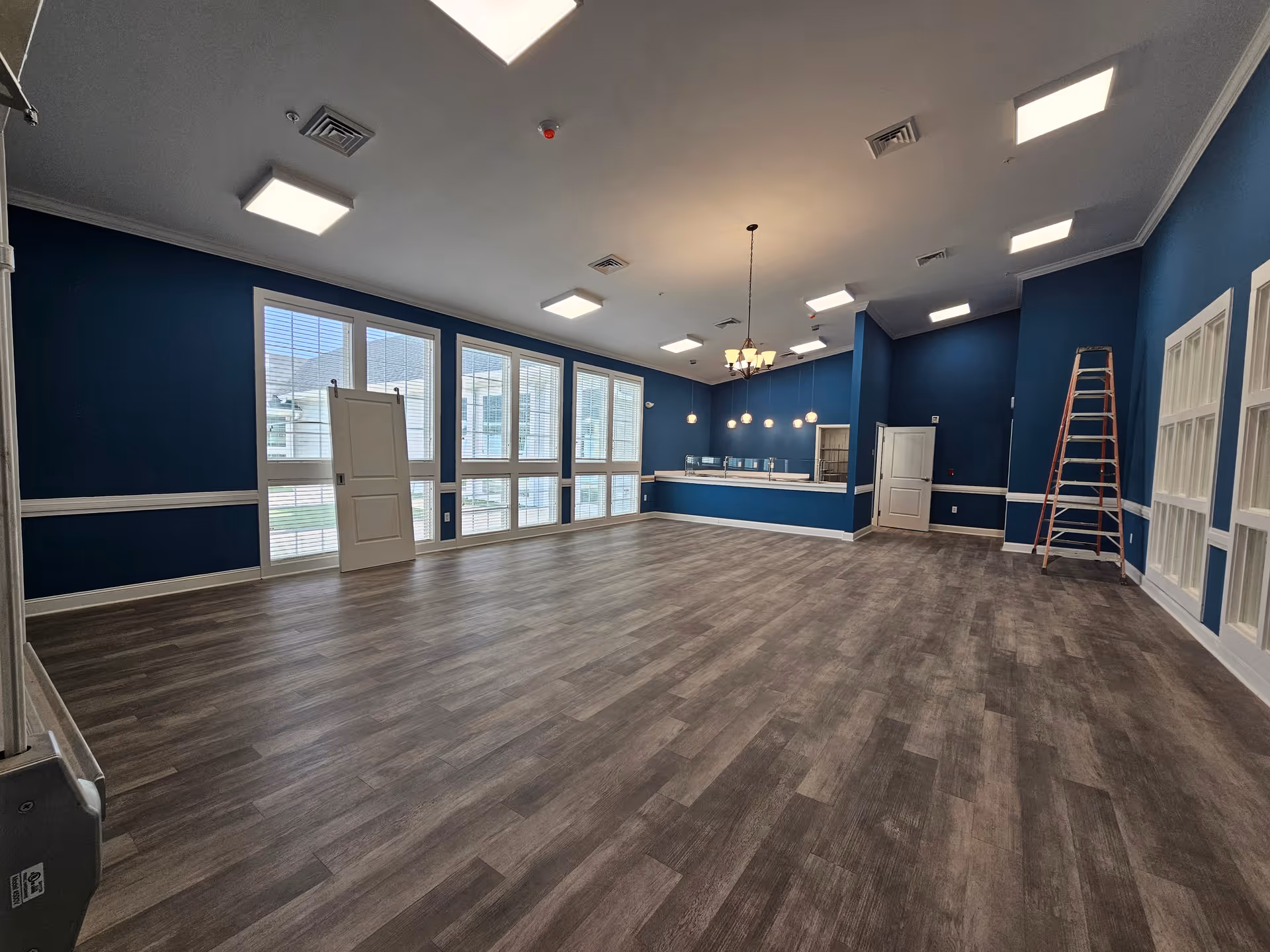 Spacious empty multipurpose room with dark blue walls, large windows, hanging lights and a service counter, with a ladder against one wall.