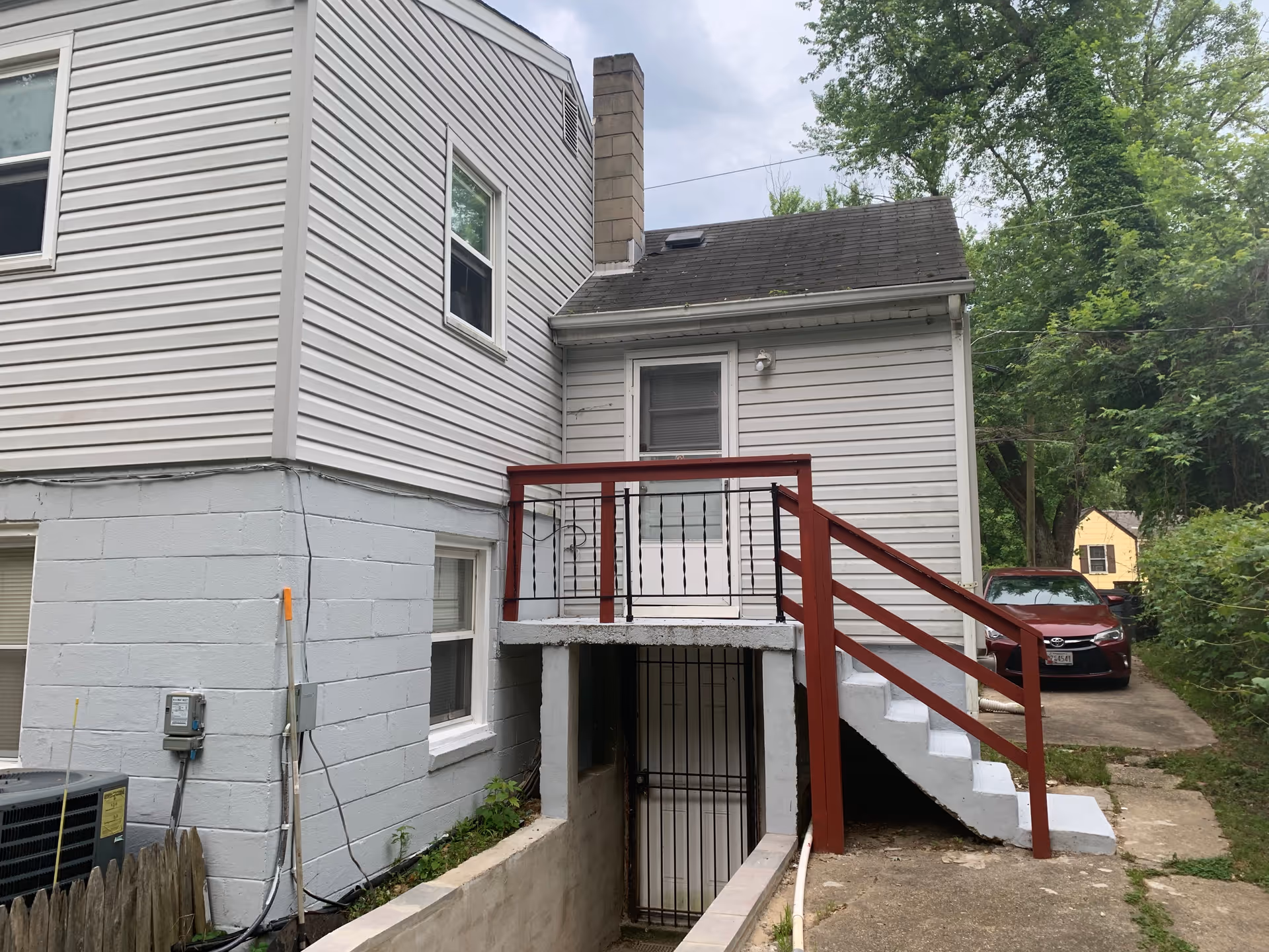 Rear exterior view of a two-story house with white siding and a gray concrete foundation. There is a small elevated porch with red railings and stairs leading down to a gated basement door. A red car is parked on a driveway to the right, and there are trees and greenery surrounding the area.