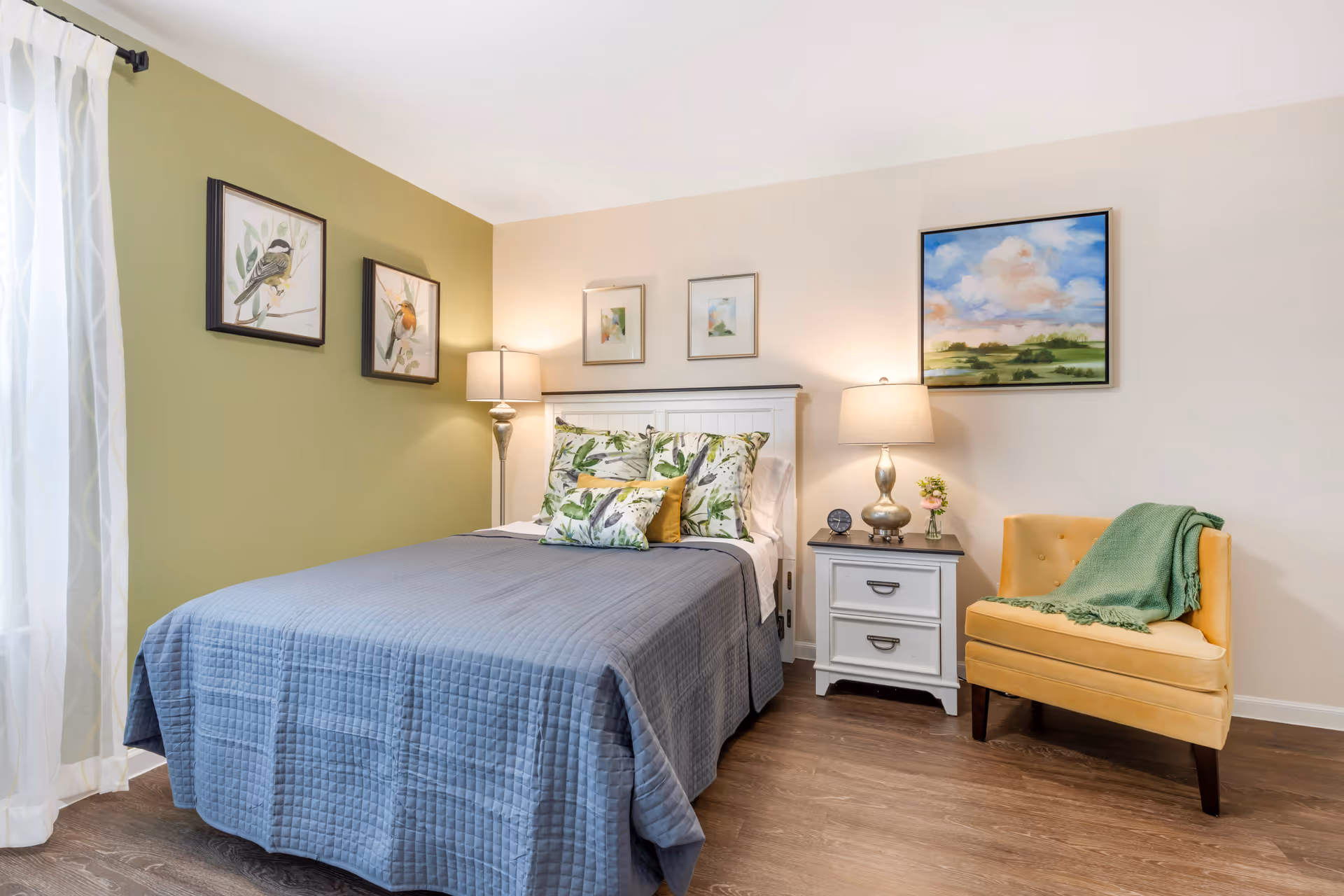 Well-lit bedroom with a bed dressed in a gray quilt, green accent wall, bedside table and lamp, framed artwork, and a yellow armchair.
