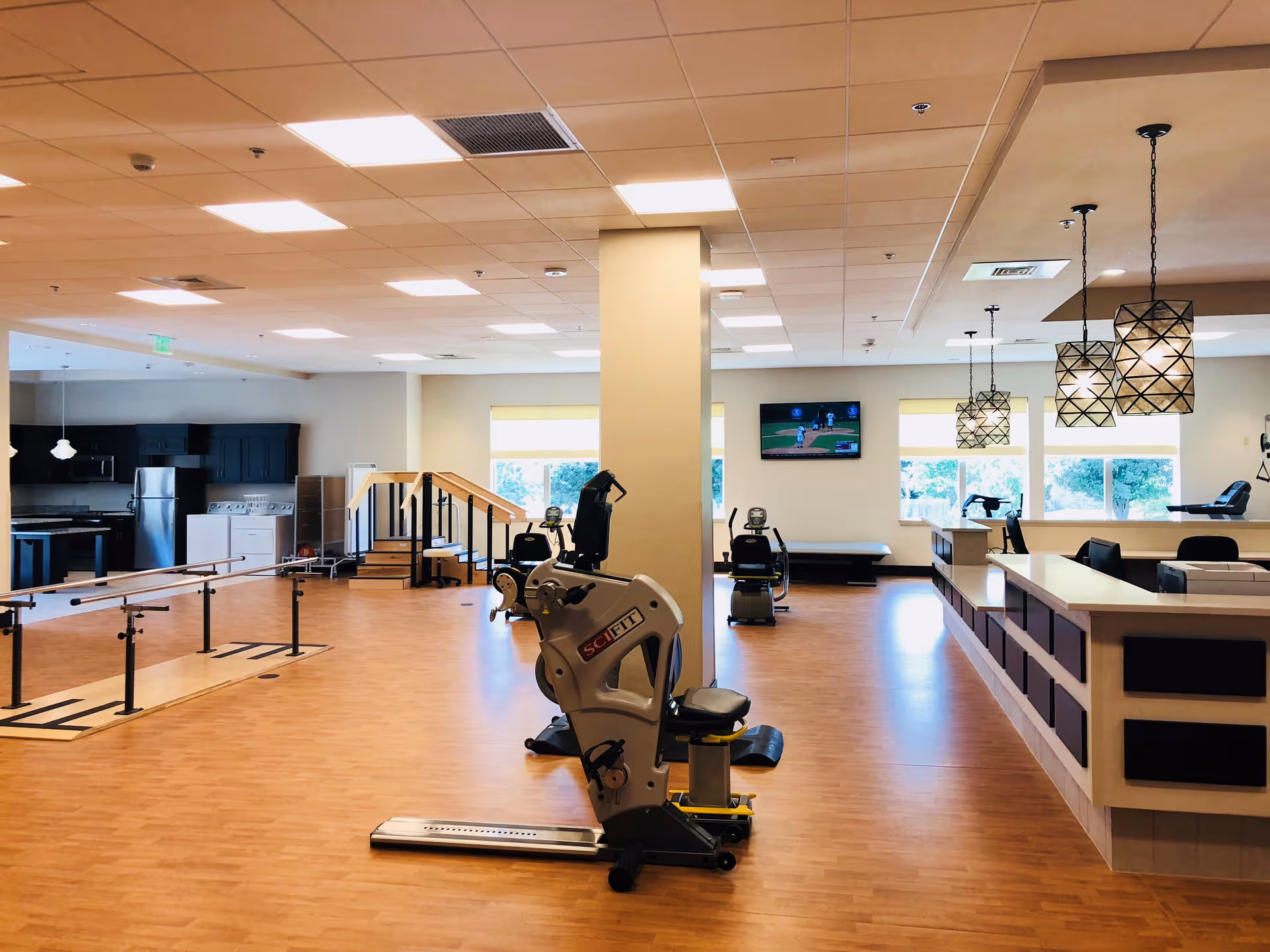 Bright, open rehabilitation gym with parallel bars, exercise machines, a practice staircase, kitchenette area and a reception counter.