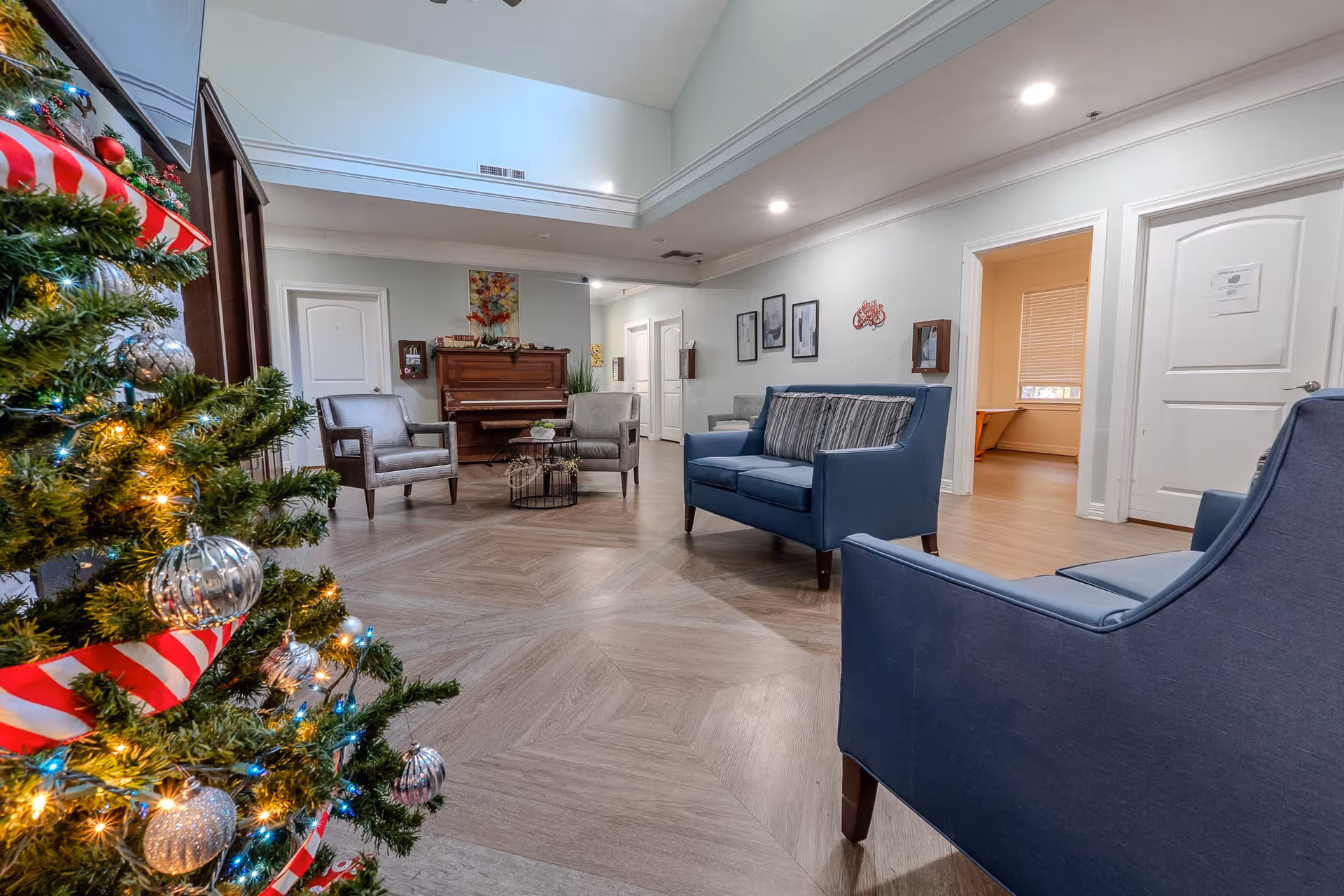 A cozy senior living common area decorated for Christmas with a Christmas tree adorned with silver ornaments and red-and-white ribbon. The room features blue upholstered chairs and a loveseat, two gray armchairs, a wooden piano against the wall, and framed artwork. The space has wood-patterned flooring and white walls with recessed lighting.