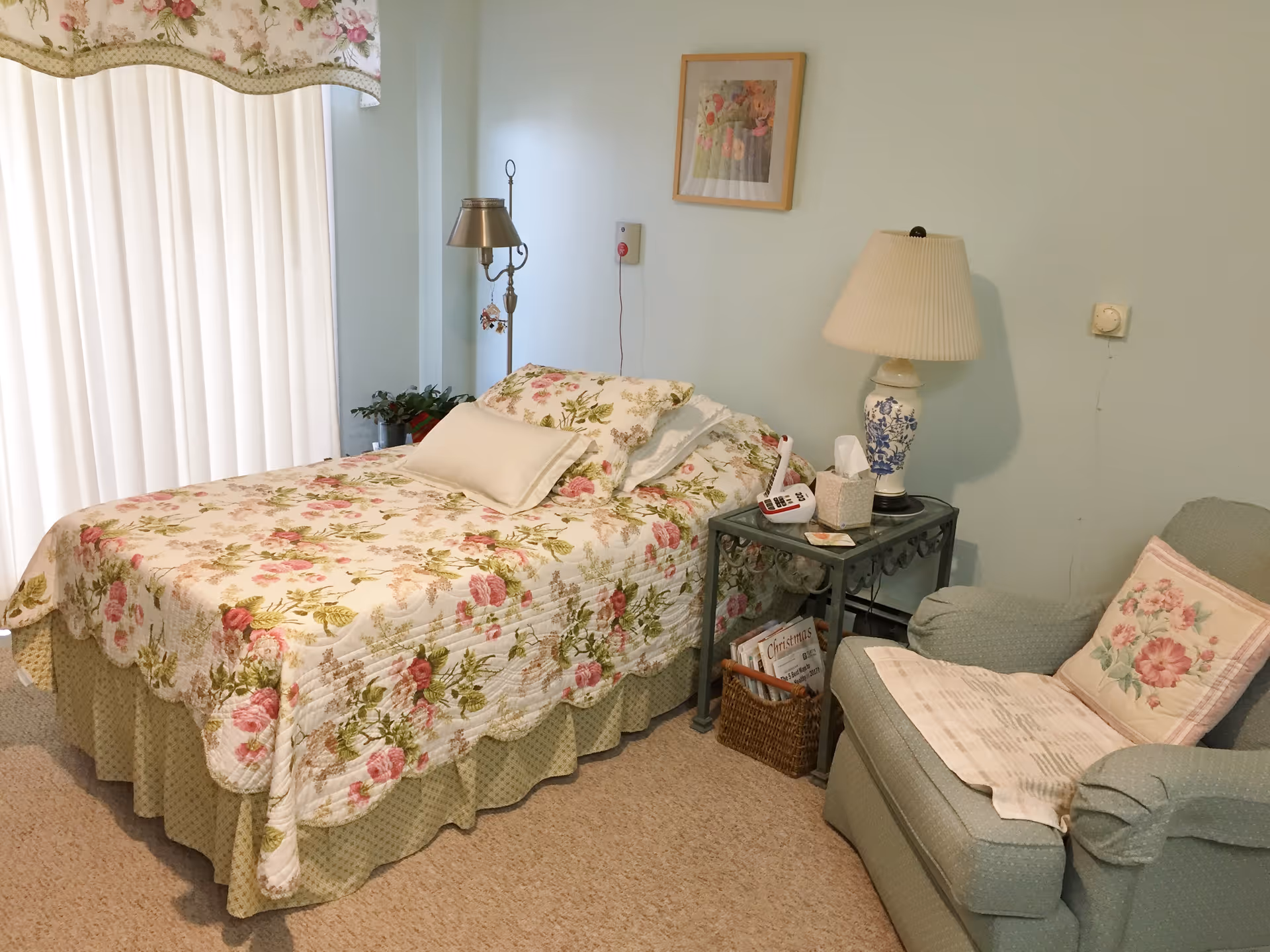 A cozy bedroom with a bed covered in floral bedding, a matching floral valance over a window with vertical blinds, a bedside table with a lamp, tissue box, and remote control, and a comfortable armchair with a floral pillow and a blanket.