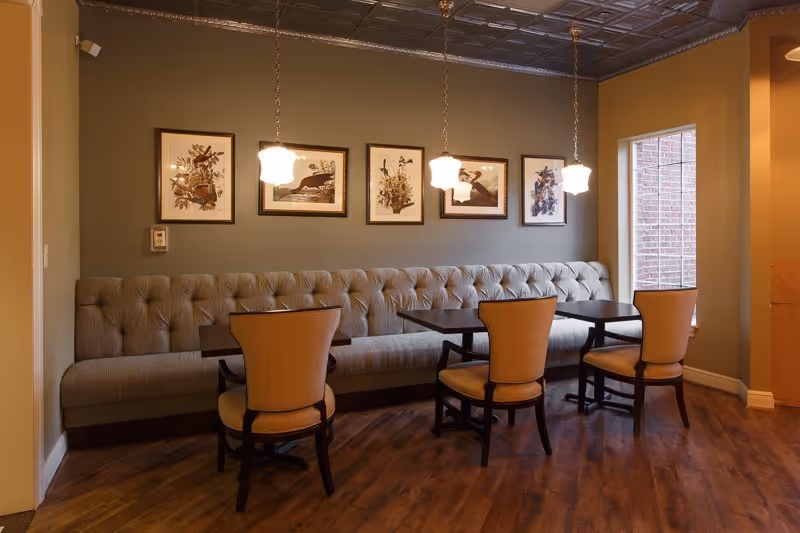 A cozy dining area with a long tufted bench along the wall and four wooden chairs with cushioned seats around two dark wooden tables. Above the bench are five framed botanical prints, and three pendant lights hang from the ceiling. A window on the right side lets in natural light, and the floor is wooden.