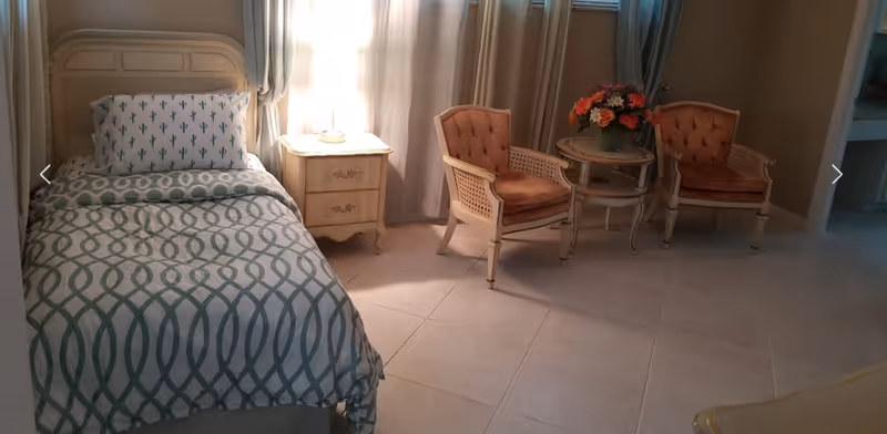 A cozy bedroom in a senior living facility with a bed, seating area, and flowers.