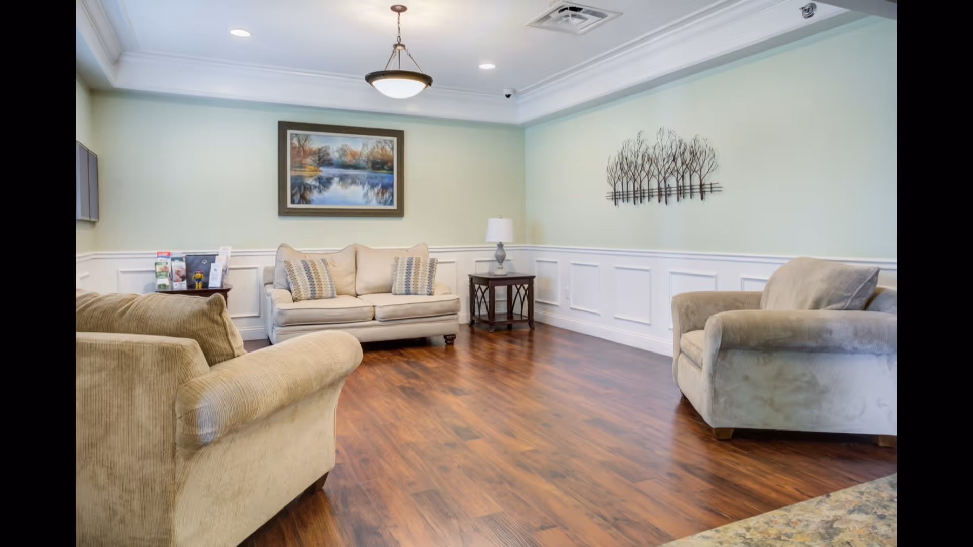 Comfortable memory-care lounge with a sofa and armchairs on a hardwood floor, wall art, and a ceiling light.