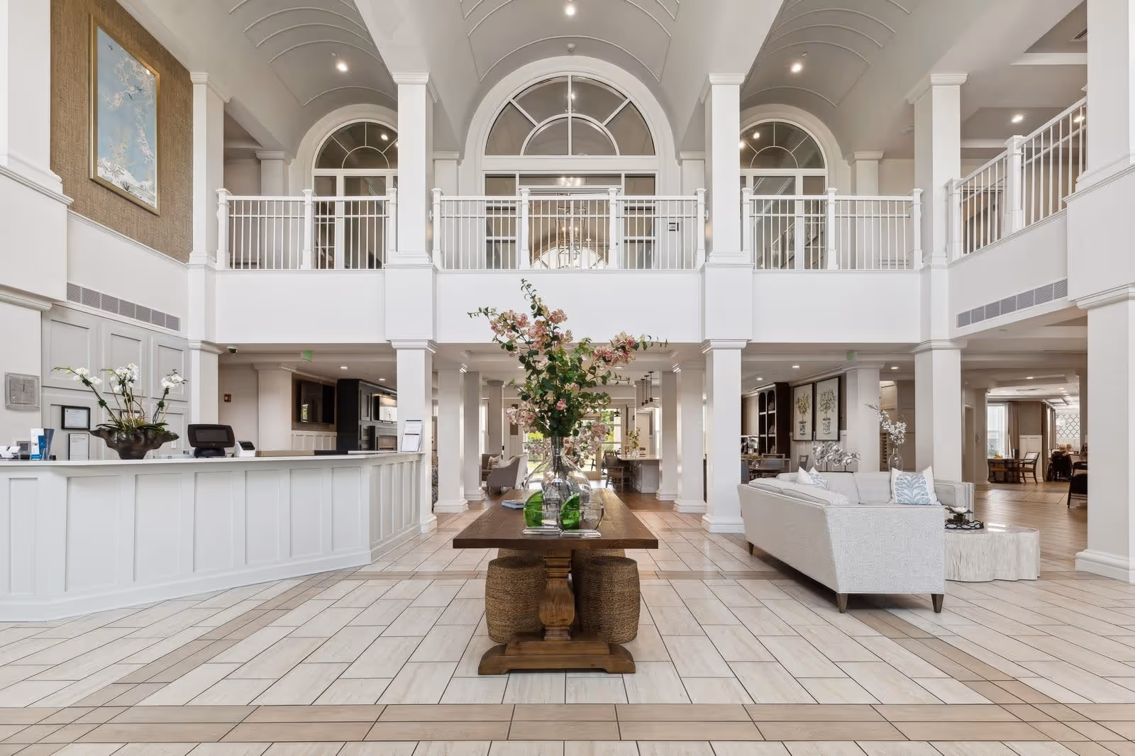 Spacious and elegant senior living facility lobby with high vaulted ceilings, white pillars, a central wooden table adorned with floral arrangements, a white reception desk on the left, and a comfortable seating area with a light-colored sofa on the right. The area is well-lit with natural light coming through large arched windows on the upper level.