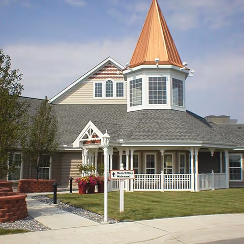 Front exterior of a senior living facility featuring a turret with a copper roof, a wraparound porch, and a sign pointing to the main office.