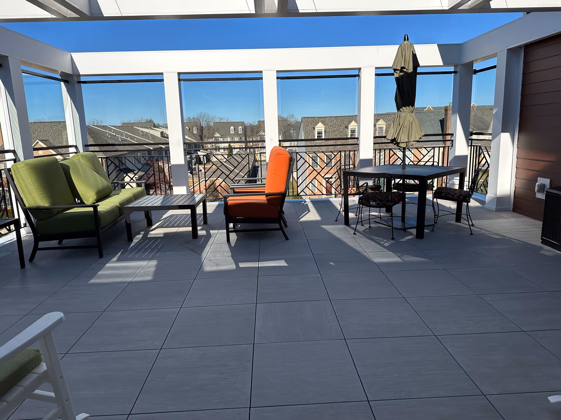 A bright terrace area with green and orange seating, a table, and an umbrella, surrounded by large windows showcasing a clear blue sky and rooftops in the background.