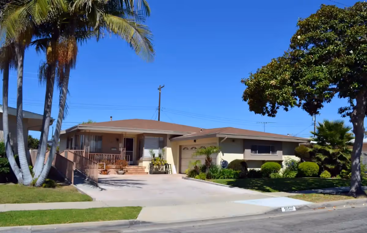 Single-story residential building with a driveway, garage, and front porch. The house is surrounded by green grass, palm trees, and other shrubs under a clear blue sky.