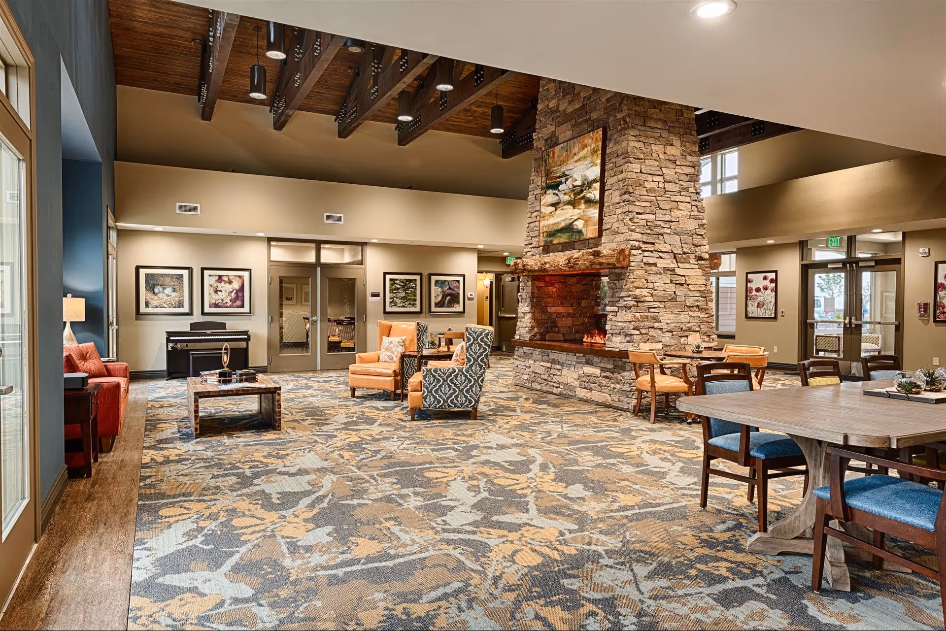 A spacious senior living facility common area with a large stone fireplace in the center. The room features a patterned carpet, wooden ceiling beams, and various seating options including armchairs, sofas, and dining tables with chairs. Artwork decorates the walls, and large windows allow natural light to enter.