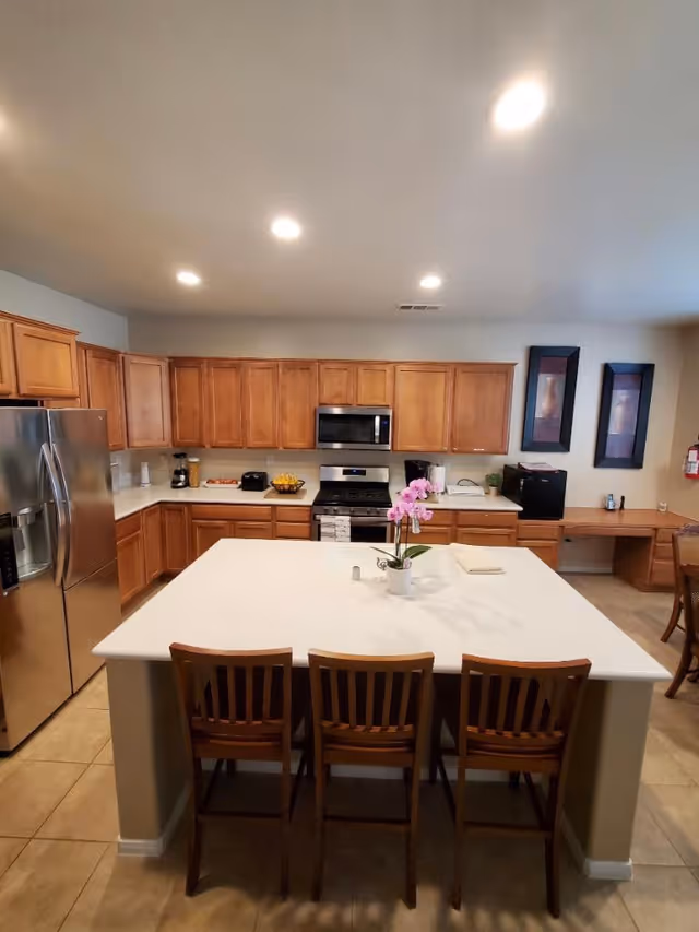 Bright kitchen with a large white island and three wooden bar stools, stainless steel refrigerator and stove, and wood cabinets.