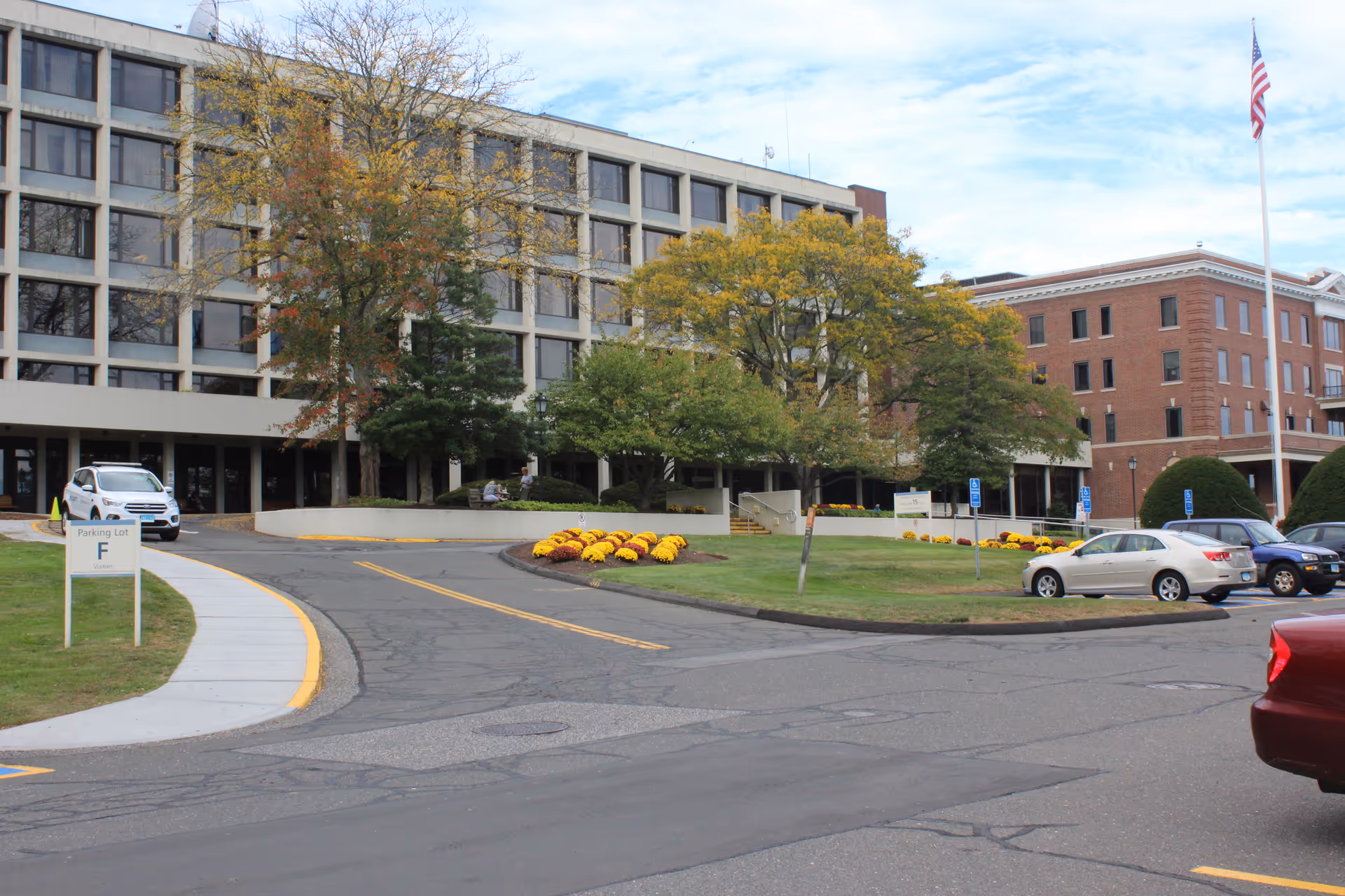 Front exterior of a multi-story health center with a driveway, landscaped flowerbeds, parked cars, and an American flag.