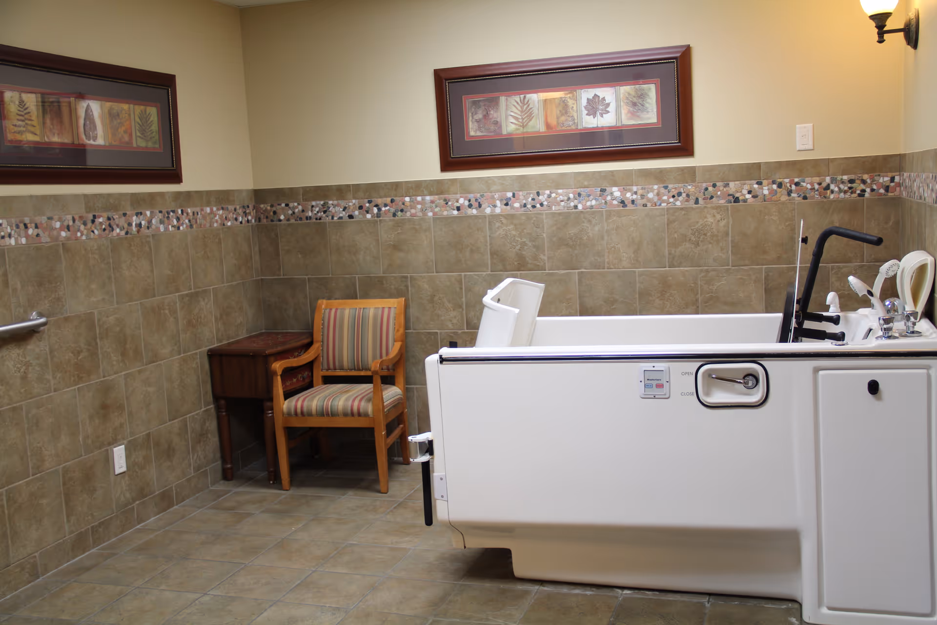 A bathroom with beige tiled walls and floor, featuring a white walk-in bathtub with safety handles and a faucet. There is a wooden chair with striped upholstery and a small wooden table in the corner. Two framed pictures with leaf designs hang on the walls, and a wall-mounted light fixture is visible.
