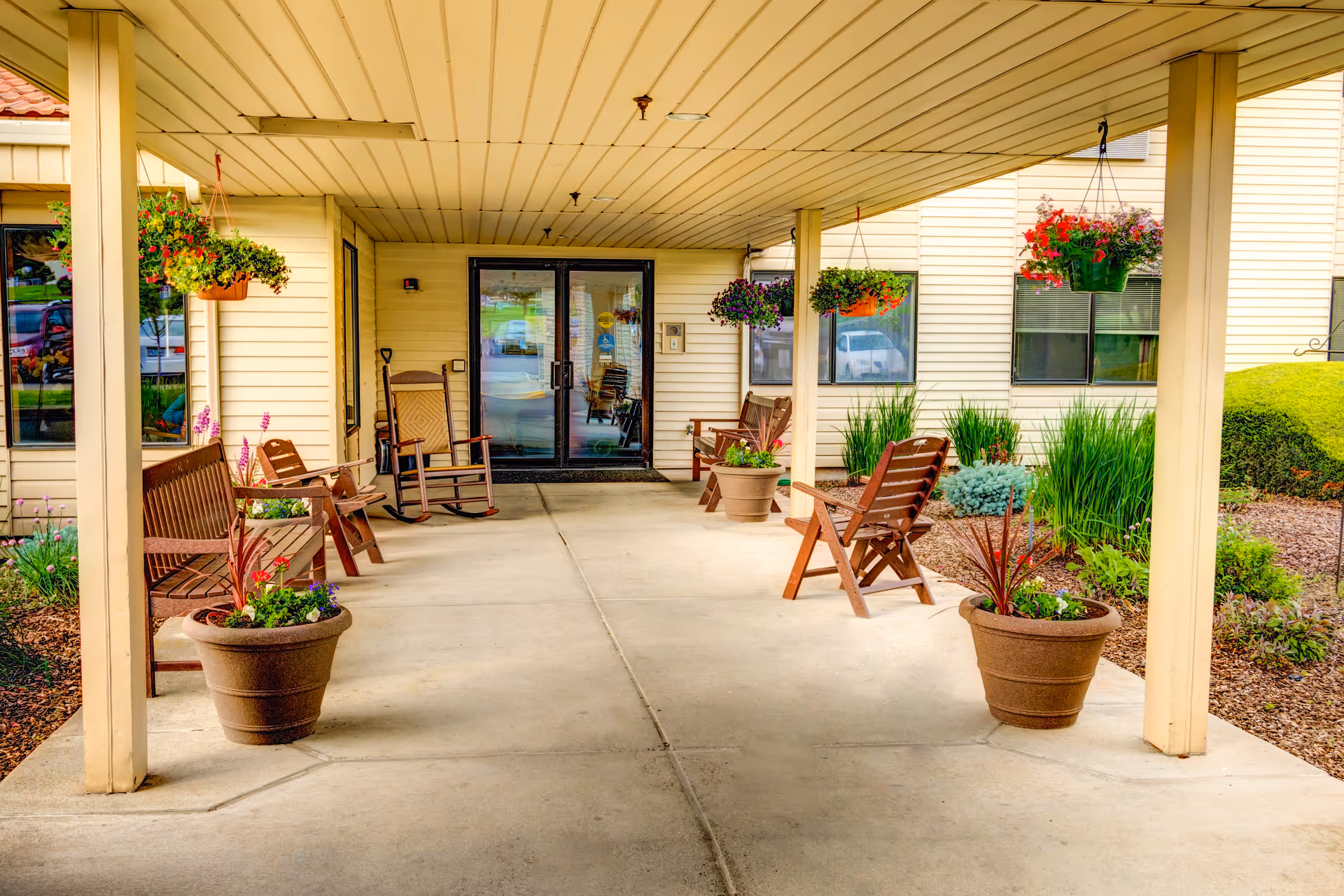 Covered entrance patio of a retirement residence with wooden chairs, rocking chairs, potted plants and hanging flower baskets in front of glass double doors.