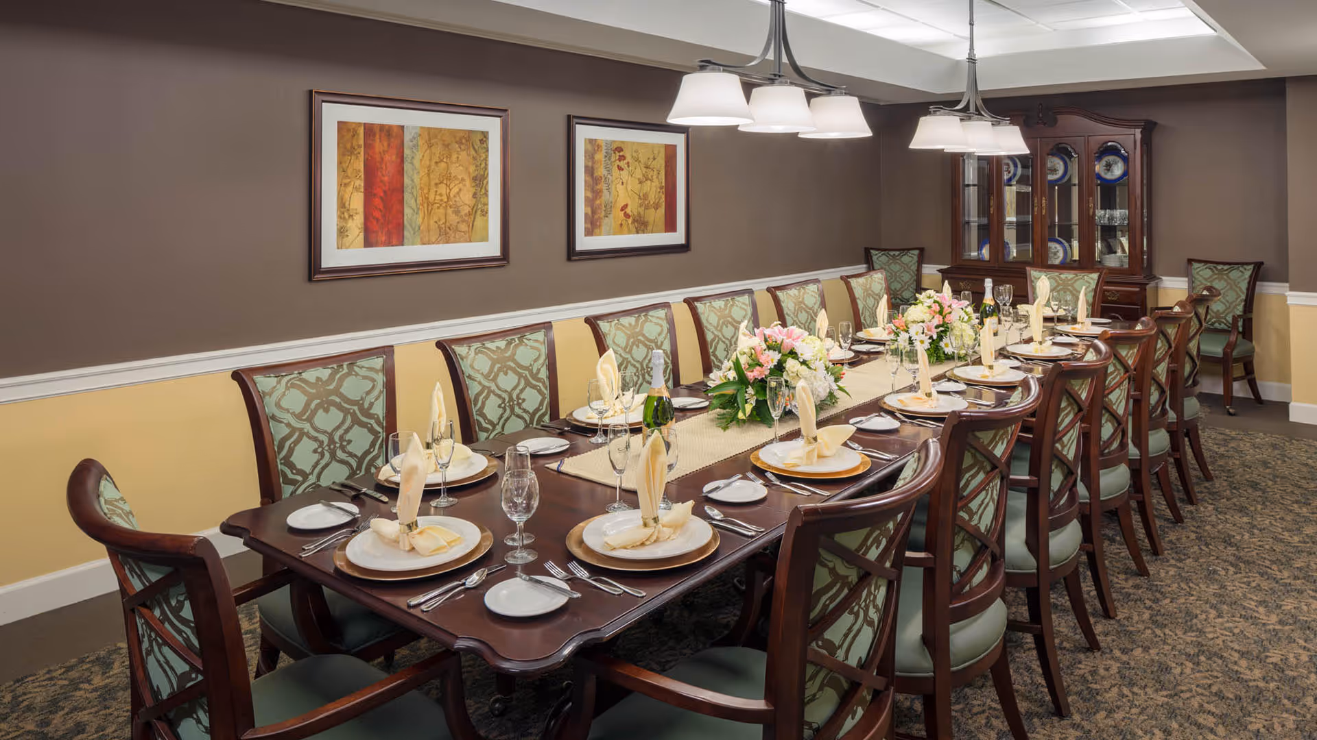 A formal dining room with a long wooden table set for a meal. The table is arranged with plates, napkins folded in an upright design, glasses, and silverware. There are floral centerpieces and a bottle of champagne on the table. The room has patterned carpet, beige and brown walls with two framed artworks, and a wooden china cabinet displaying plates in the background. Several upholstered chairs with wooden frames surround the table.