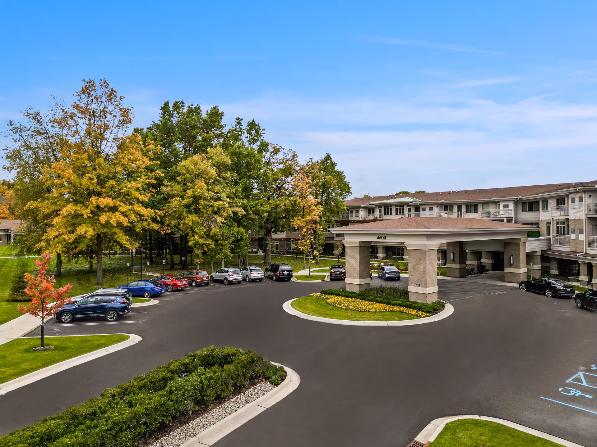 Exterior view of a senior living facility named All Seasons Ann Arbor with a circular driveway, parked cars, landscaped greenery, and trees with autumn foliage under a partly cloudy sky.