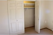 Empty bedroom closet with white bi-fold doors partially open, beige carpeted floor, and light yellow walls.
