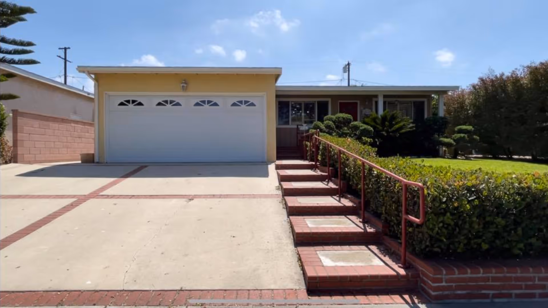 Front exterior view of a single-story house with a two-car garage, a concrete driveway with brick accents, a set of brick stairs with a red handrail leading to the front door, and well-maintained bushes and lawn on the right side under a clear blue sky.