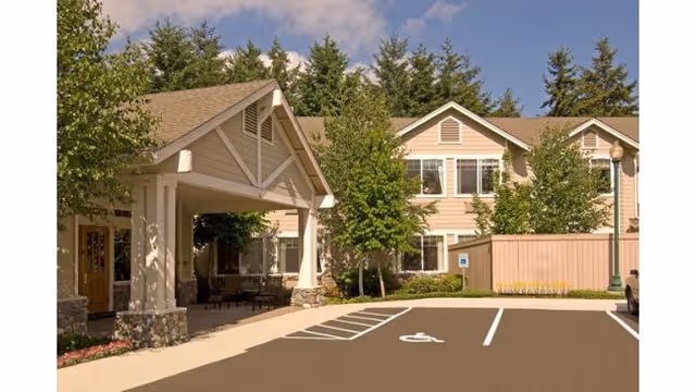 Exterior view of a senior living facility with a covered entrance, parking spaces including a handicapped spot, trees, and a two-story building with beige siding and white trim under a blue sky.