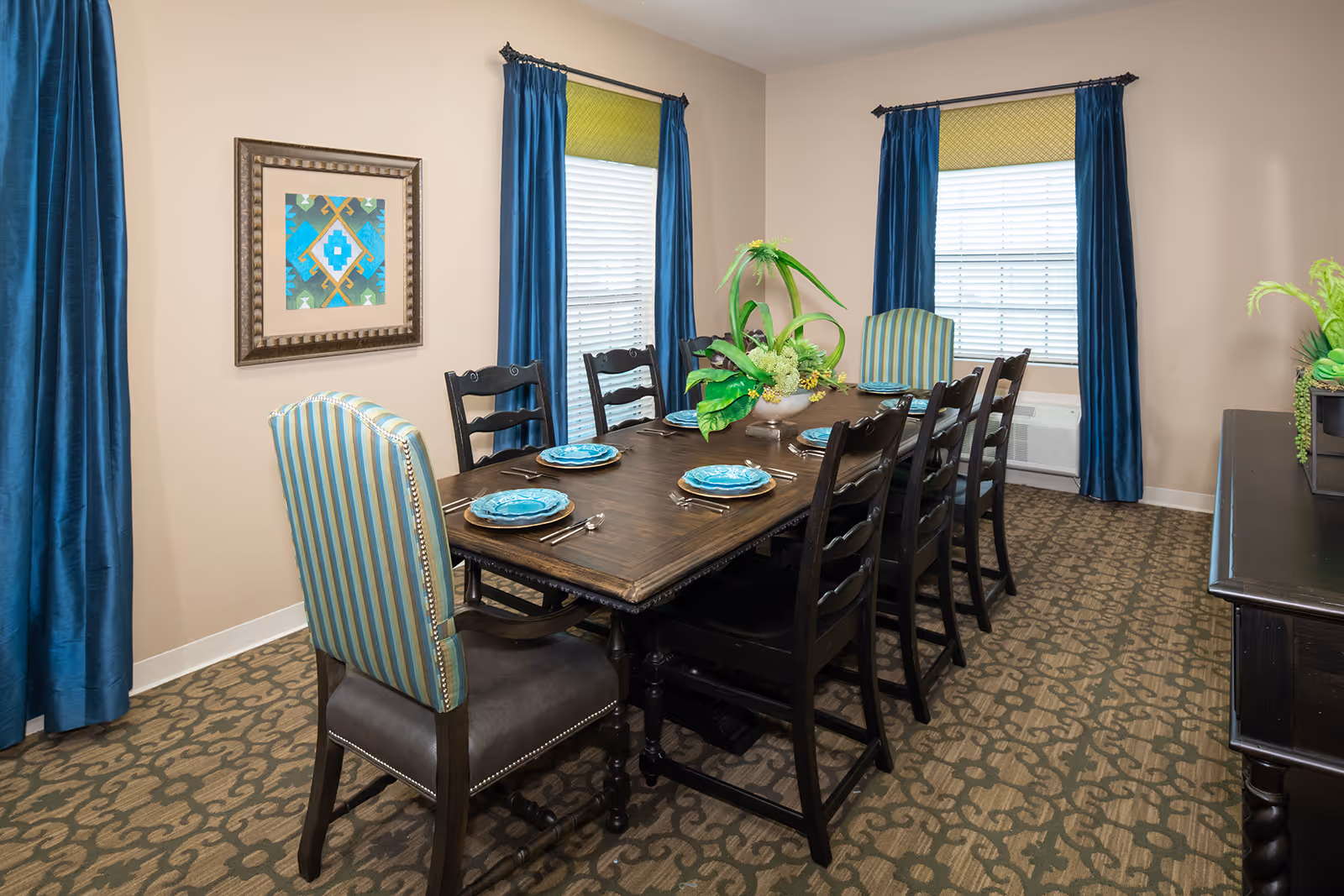A dining room with a dark wooden table set for six people with blue plates and silverware. The room has two windows with blue curtains and green valances. There are six black wooden chairs and two striped upholstered chairs at the ends of the table. A decorative plant centerpiece is on the table, and a framed geometric artwork hangs on the beige wall.